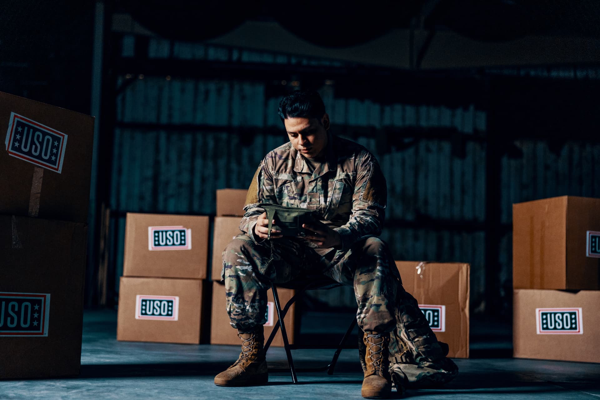 A soldier sitting quietly among USO care-package boxes in a dim warehouse.