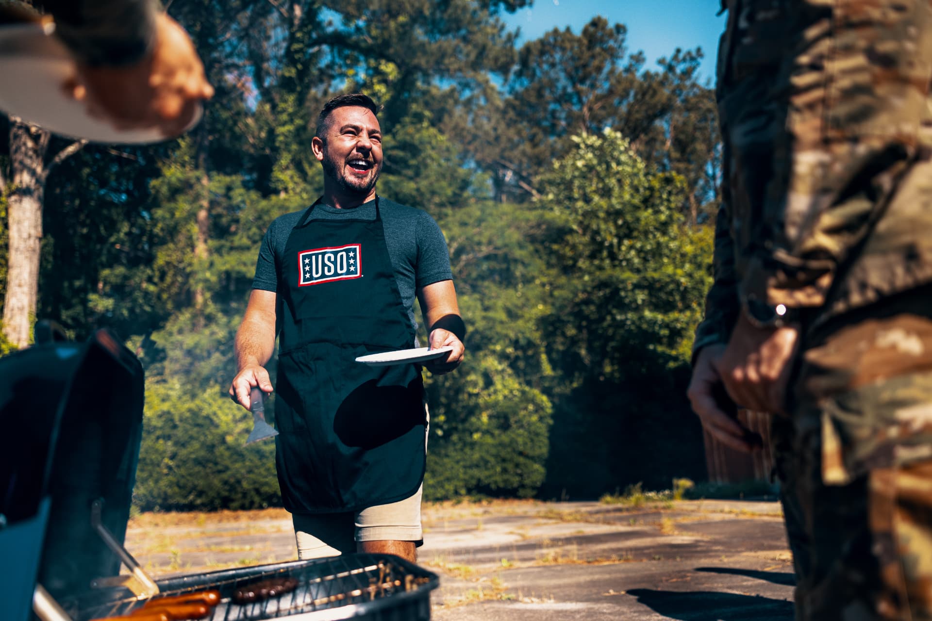 A USO staff member in an apron laughing while grilling at a summer cookout for troops.