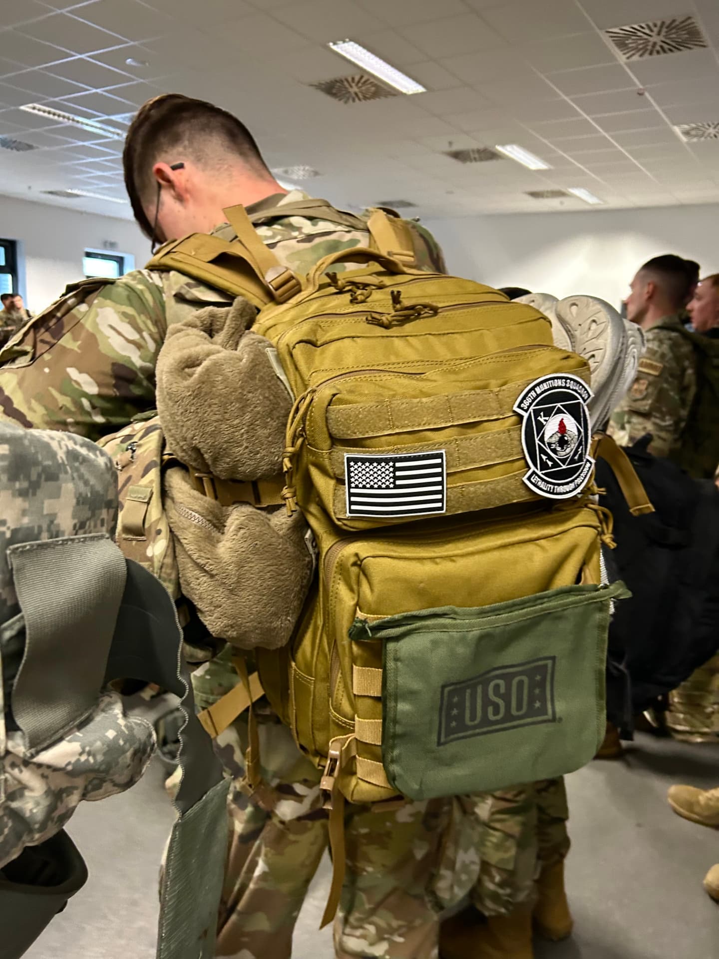 A green USO care pack clipped to an airman’s tan rucksack before a movement.