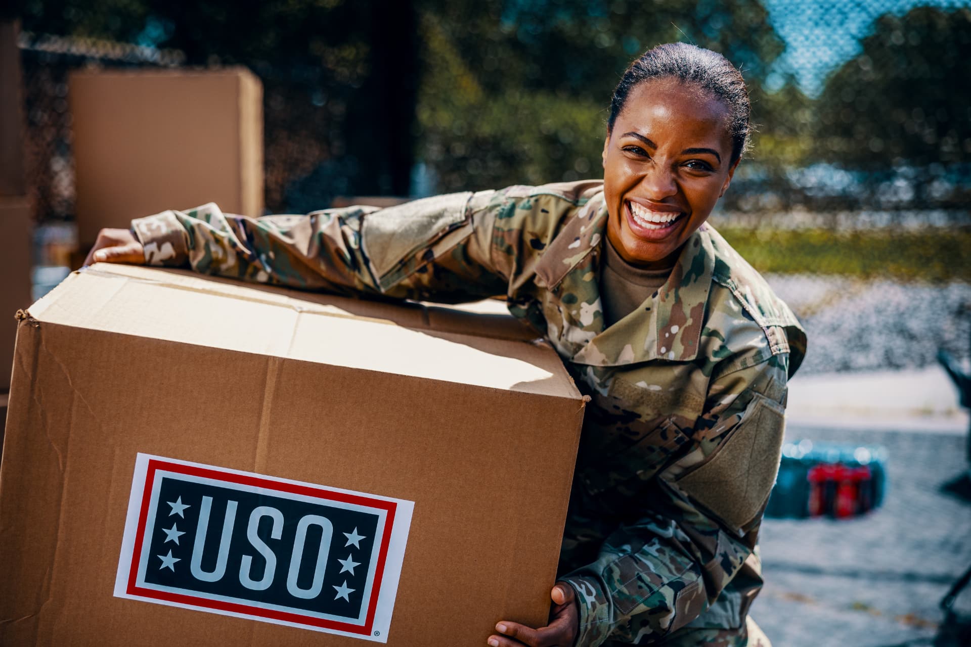 A smiling soldier holding a USO care pack at an outdoor event.