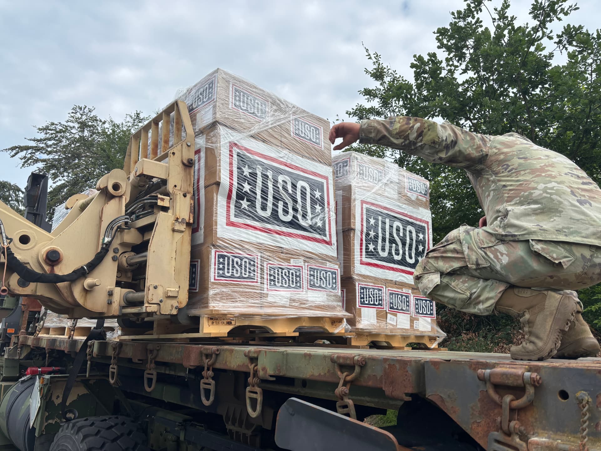 A soldier steadying shrink-wrapped USO pallets loaded on a military flatbed truck.