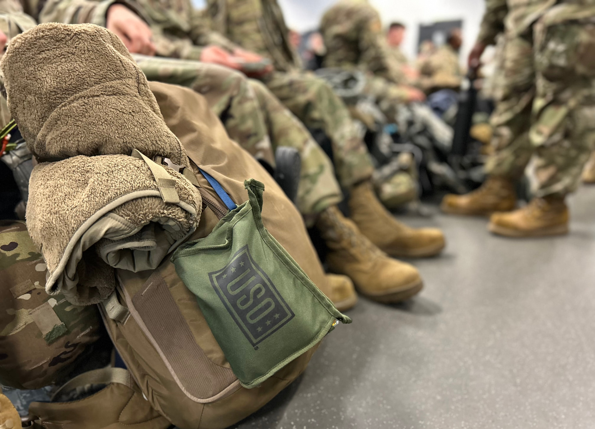 A green USO care pack clipped to a packed rucksack as troops wait to move out.