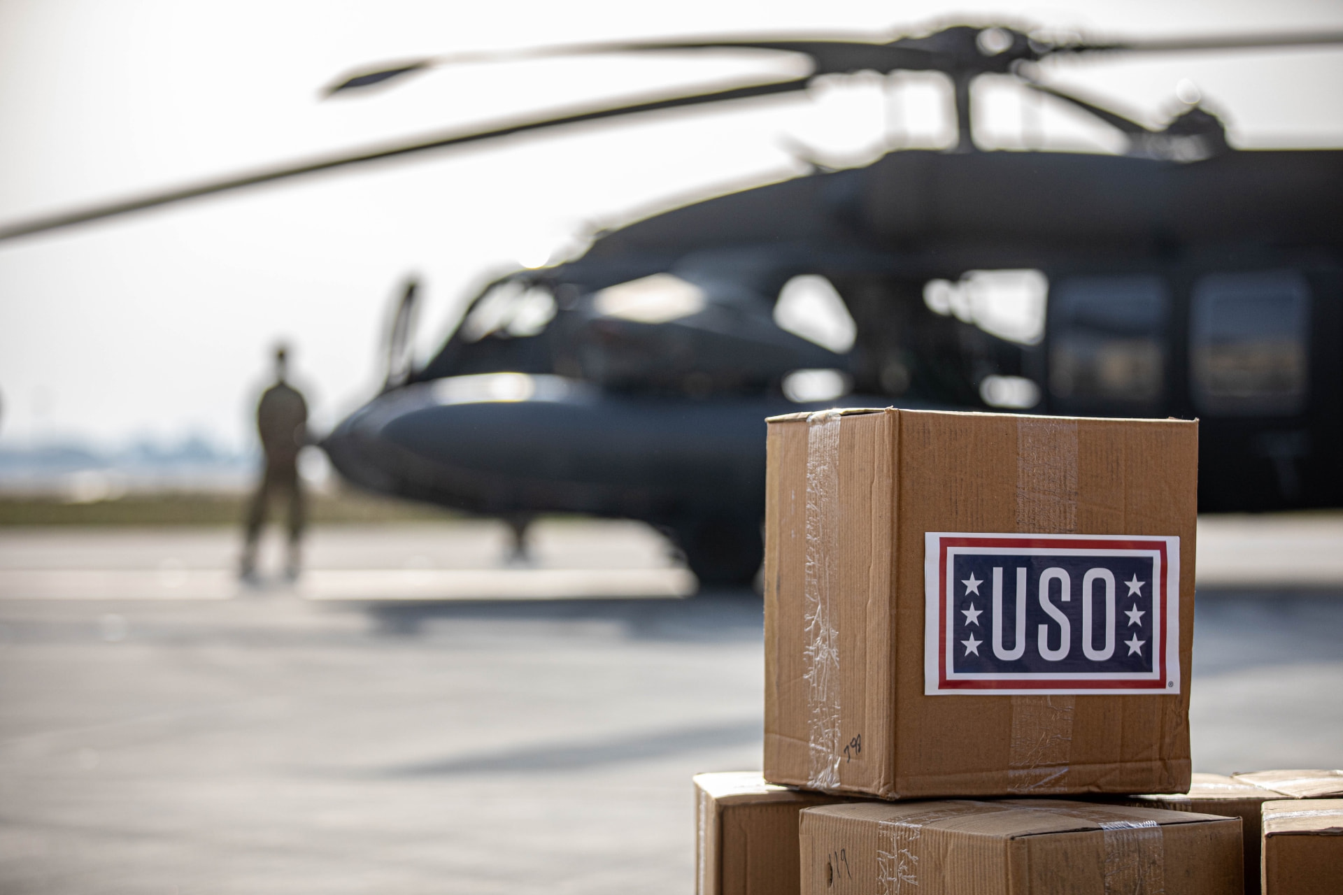 USO care package boxes stacked on a flight line in front of a military helicopter.