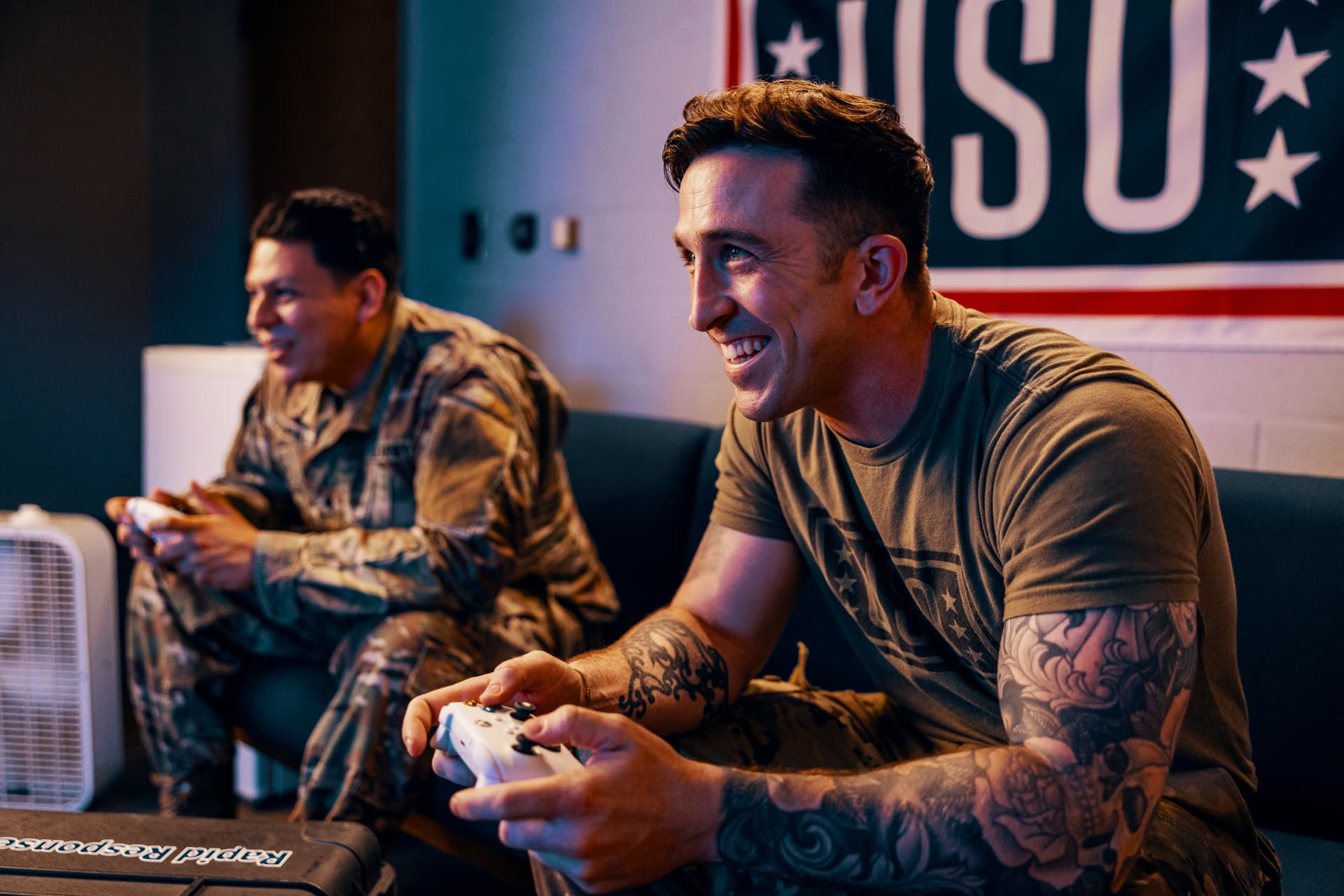 Two service members laughing while playing video games with controllers under a USO banner.