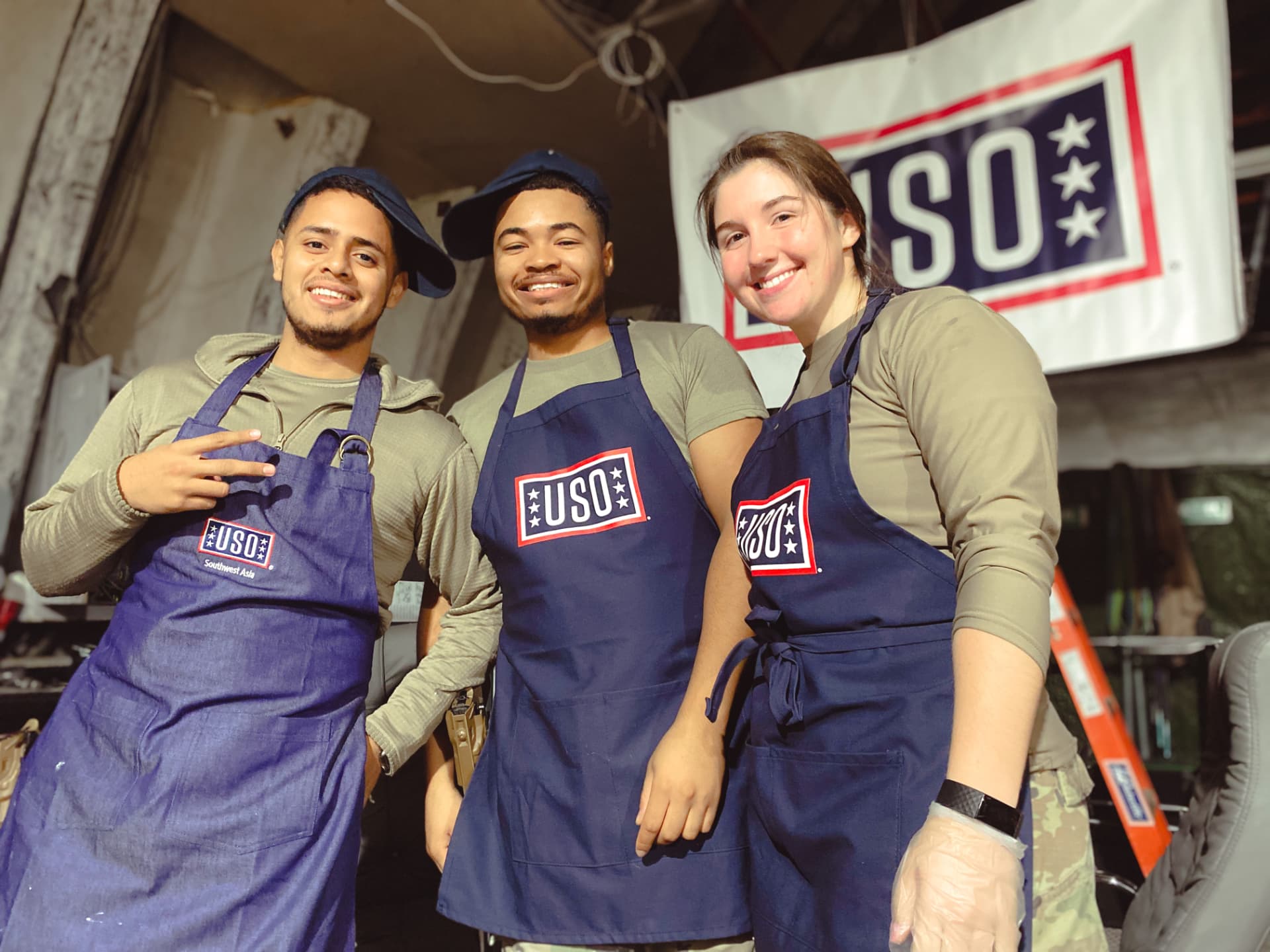 Three smiling USO volunteers in branded aprons in front of a USO banner.