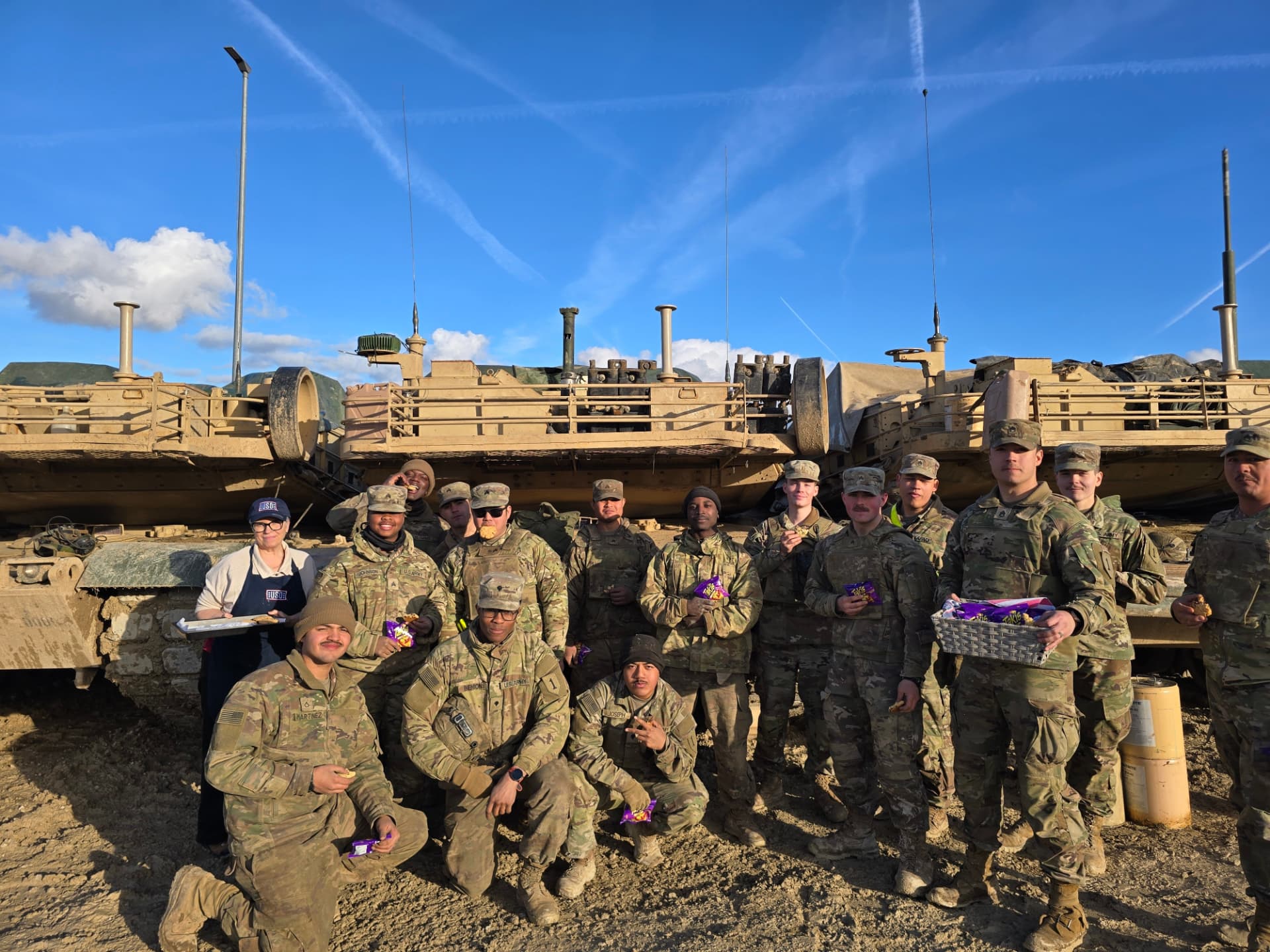 USO volunteers handing out snacks to a group of U.S. Army soldiers beside armored vehicles at a training area in Germany.