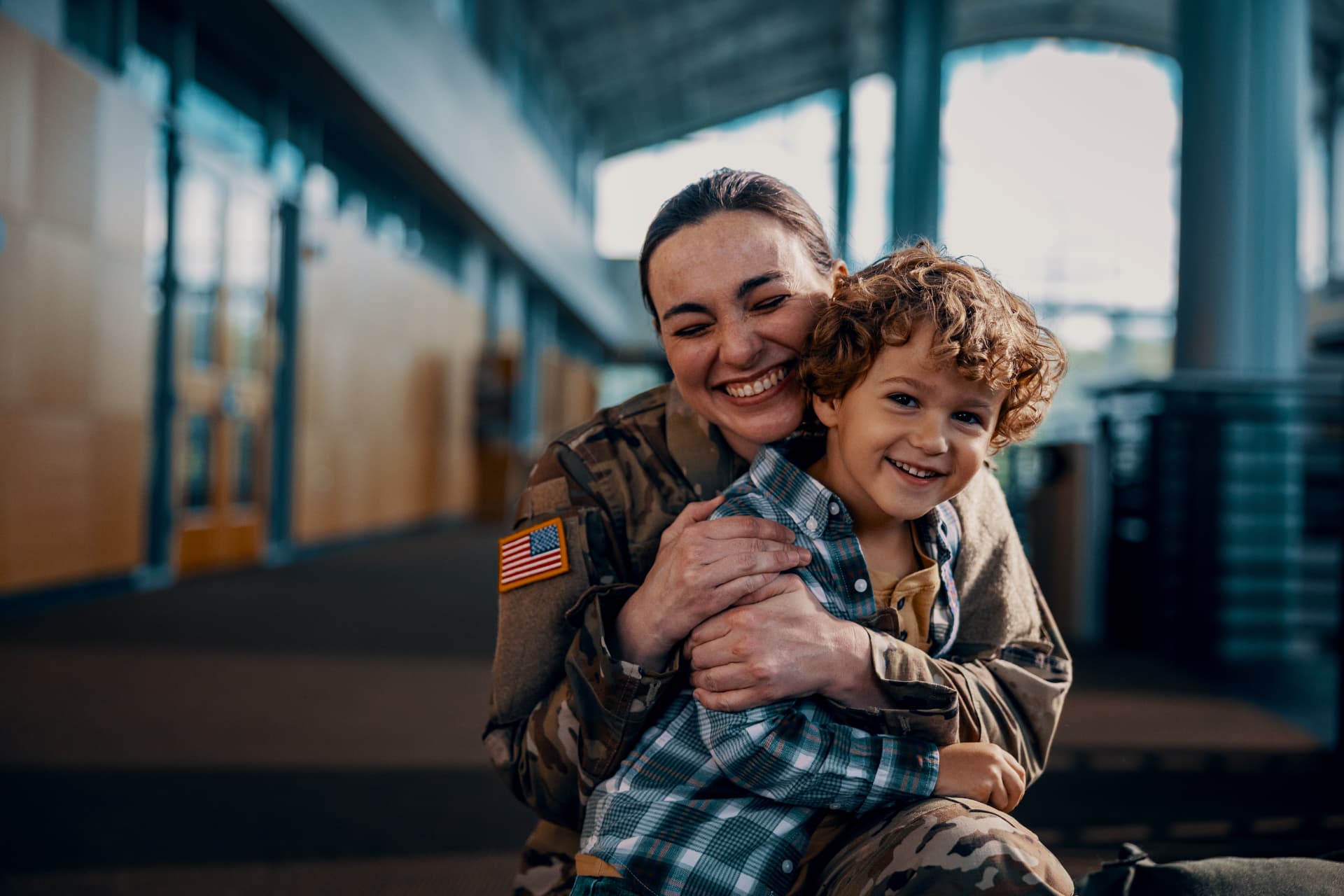 A soldier in uniform hugging her young son at an airport terminal, both smiling.