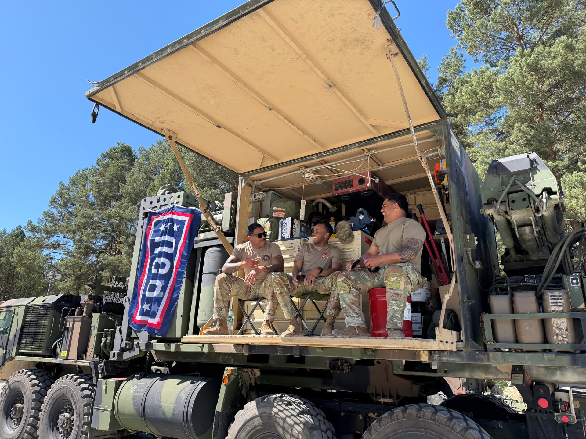 Three soldiers laughing on the back of a truck beside a USO banner during an exercise.